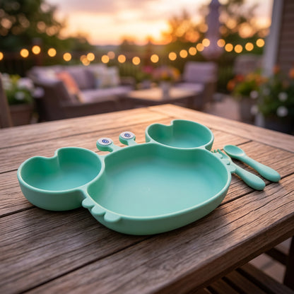 A mint green silicone baby feeding tray set on a wooden table, with a blurred outdoor patio background at sunset.