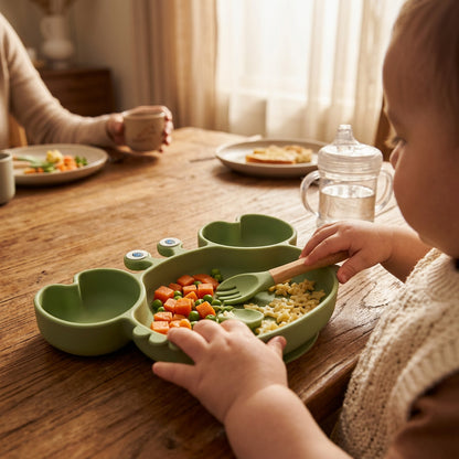 A light green silicone baby feeding tray shaped like a frog, with two heart-shaped side bowls, two small eye details, and two wooden utensils (a fork and a spoon) placed inside. The set is photographed on a solid pale peach-beige background.