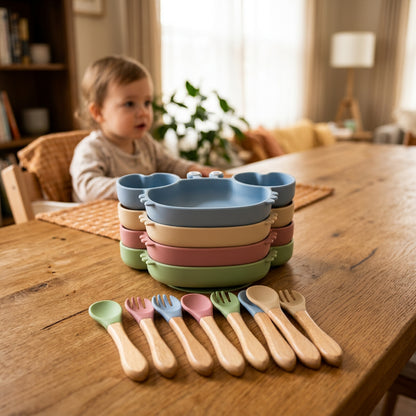 A set of silicone baby feeding accessories on a warm beige background, including a large and small plate in a light blue mouse-head shape, a stack of colorful plate lids, and a matching wooden utensil set with spoons, forks, and spatulas.