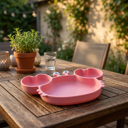 A pink silicone baby feeding tray with a cartoon face and small blue eyes, shown on a wooden outdoor table with a blurred garden background.